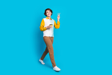 Poster - Photo of positive girl marketer walking hold latte cup make v sign greeting isolated blue color background