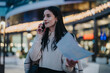 © qunica.com - A young woman talking on her phone and holding a paper in a city environment during the evening, with lights and urban architecture in the background, portraying modern business and connectivity.