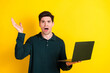 © deagreez - Young man gesturing surprisedly while holding a laptop against a vibrant yellow background in casual green attire