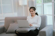 © Dara-Sroi - Asian woman living lifestyle wearing wireless headphones staring at laptop screen and communicating with clients freely on sofa at home.