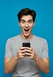 © AlfaSmart - Surprised young man using smartphone on blue background for social media engagement