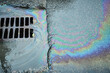© AleksFil - Water droplets create a vibrant oil slick on the pavement, displaying rainbow colors. A drain collects falling rainwater as nearby puddles form.