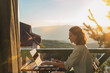 © olezzo - Young woman sitting on balcony with view of mountains and sunset working using laptop