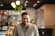 © Wavebreak Media - Mid adult man smiling at camera seated in modern cafe amid wooden decor and lush plants