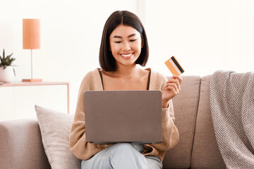 Poster - Online Shopping And Delivery Concept. Happy asian woman making purchases on internet with laptop, holding and showing credit card to pay. Smiling lady using computer sitting on sofa, copy space