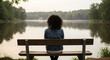 © Gabriel - A woman sitting alone on a wooden bench beside a quiet lake, reflecting trees and a pale sky, with soft wind gently blowing her hair.