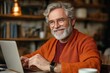 © 宜然 左 - Smiling elderly man with gray hair and beard wearing glasses and orange sweater working on a laptop in a cozy, warmly lit room