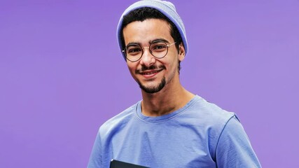 Aufkleber - Medium zoom AI animated portrait of smiling young Middle Eastern male student or freelancer in blue tshirt and beanie, holding laptop, posing in studio on purple background, looking at camera