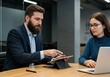 © Vooglo - Man pointing at tablet screen while woman looks at laptop during business meeting
