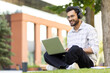 © Tetiana - A young student wearing headphones smiles while working on his laptop outdoors on the grass.