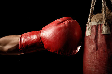 Fototapete - close-up of clenched fists, one wrapped in dark textured gloves, in front of a worn red punching bag
