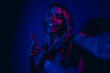 © deagreez - Young stylish woman posing in vibrant blue and pink neon lighting, expressing confidence and fun in a modern nightlife setting
