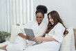© Ekkasit A Siam - Two teenage girls in white spa robes relaxing on bed smiling and enjoying tablet together, African American and mixed Caucasian Asian friends bonding through digital leisure and feminine lifestyle