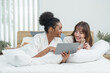 © Ekkasit A Siam - Two teenage girls lying on bed in white robes looking at tablet together, African American and mixed Caucasian Asian friends sharing joyful skincare routine and cozy modern lifestyle moments