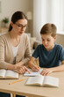 © LK  - A female teacher helps a boy do his homework at a table with books in a cozy room. The concept of tutoring and home education.