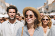© Nataliia - A cheerful close-up photograph capturing a woman wearing a straw hat and sunglasses, smiling brightly while exploring a sunny city street filled with blurry people and buildings in the background