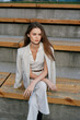 © SHOTPRIME STUDIO - stylish woman in light suit sitting on wooden bleachers outdoors with calm expression, casual fashion, spring season, natural light, modern casual style