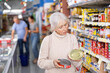 © JackF - Focused interested elderly woman standing in grocery store aisle carefully reading nutritional information on tin cans while choosing right canned food..