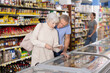 © JackF - Positive senior couple, man and woman, shopping together in supermarket, looking at goods in glass showcase and discussing