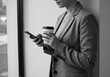 © Vooglo - Woman in blazer using smartphone while holding a coffee cup near a window in monochrome