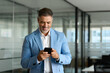 © insta_photos - Smiling 50 year old smiling businessman professional holding smartphone looking at camera at work. Happy middle aged business man investor wearing suit standing in office using mobile phone.