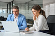 © insta_photos - Professional mature business man and woman leaders colleagues talking working on computer at workplace. Two busy professional executives company team working in corporate office using pc laptop.