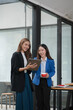 © Thitisak - Two  Asian businesswomen holding iPads, holding graph papers, standing in front of the glass, talking and consulting with each other.