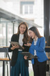 © Thitisak - Two  Asian businesswomen holding iPads, holding graph papers, standing in front of the glass, talking and consulting with each other.