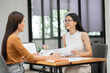 © aekachai - Two Asian businesswomen holding graph documents, sitting and talking about work, looking happy while working.