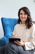 © Andrii Zastrozhnov - Smiling young hispanic female holding tablet and stylus seated on blue couch