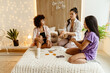 © Drobot Dean - A multiracial group of three young adult women sits on a bed indoors, wearing casual clothes and applying skincare patches while talking and spending time together in a cozy bedroom.
