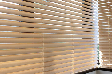 Poster - Corner windows fitted with venetian wood blinds allow soft natural light to enter the room. The warm-toned horizontal slats of half-closed wooden shutters 50mm create a cozy atmosphere.
