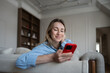© ingoodtime - Happy midlife woman lounging on cozy couch at home, scrolling through phone with joy. Relaxed interested mature female smiling reading something pleasant on smartphone resting comfortably in apartment