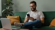 © NN AI - Caucasian man using smartphone on green sofa with laptop nearby. Remote work and digital communication concept. Home office and online connectivity. Modern lifestyle and technology integration