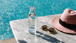 © N Joy Art  - Clear glass water bottle on marble table beside pink hat and sunglasses near blue pool on sunny day for summer lifestyle