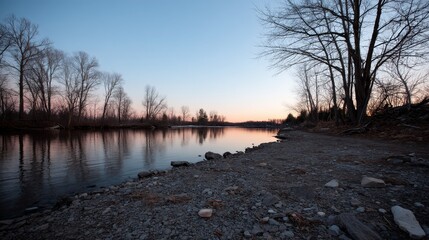 Aufkleber - A calm lake captures soft pink light from the setting sun, surrounded by bare trees and a rocky shoreline under a fading sky