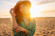 © maxbelchenko - Portrait of happy woman in straw hat walking on sunset beach. Young woman in dress enjoying sea view outdoors, feeling freedom. Active lifestyle