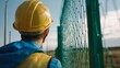 © Adi - Close-up of a construction worker wearing a yellow helmet and blue vest, looking at a green industrial gate with a metal mesh. In the background, there is an open field