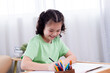 © Rakchanok - Portrait of young cheerful Asian multiethnic girl writing schoolwork or coloring book spend weekend free time at home, little kid studying homeschool learning attentive, education of elementary kid