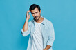 © SHOTPRIME STUDIO - Handsome man wearing light denim shirt looking at camera with playful expression while touching his hair against solid blue background. Fashion lifestyle concept.