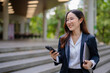 © maeching - Asian businesswoman in suit smiling holding smartphone and documents as she walks through modern business building