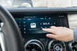 © Vladimir Razgulyaev - A closeup view of a cars modern dashboard showcasing a sleek digital display alongside well placed radio controls. The passenger controls the radio on the car's touchscreen with his finger.