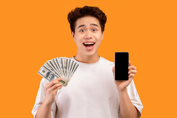 Poster - Surprised asian guy holding mobile phone with black empty screen and fan of cash money in dollar banknotes in hand, posing isolated on orange background. People and finance concept. Mock up copy space