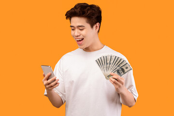 Poster - Make Money Online. Portrait of happy asian teen guy holding and using smartphone, showing bunch of money cash, standing isolated over orange studio background with free space. Cashback