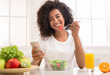 © Prostock-studio - Gadget addiction. Happy black woman texting on smartphone while eating vegetable salad at home