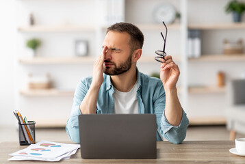 Poster - Caucasian male freelancer feeling tired, rubbing irritated eyes, sitting at desk with laptop, exhausted from online work. Young man having visual fatigue, doing business project on web