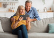 © Prostock-studio - Happy Senior Couple Using Digital Tablet Together Browsing Internet Sitting On Sofa At Home. Retired Husband And Wife Reading Online News On Computer On Weekend. Mature People And Gadgets