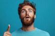 © smartcat - Surprised young man with beard looking upwards with wide open blue eyes and slightly open mouth wearing blue shirt against plain blue background