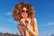 © SHOTPRIME STUDIO - Smiling woman with curly hair wearing heart shaped sunglasses holding a slice of watermelon at the beach Bright blue sky in the background
