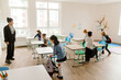 © Drobot Dean - A multiracial group of school-aged children enters a brightly lit classroom with backpacks, interacting near desks. A female Black teacher in business attire watches them with a smile.
